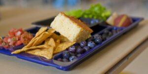 A blue school lunch tray showing corn chips and salsa, cornbread, fresh blueberries, and salad greens.