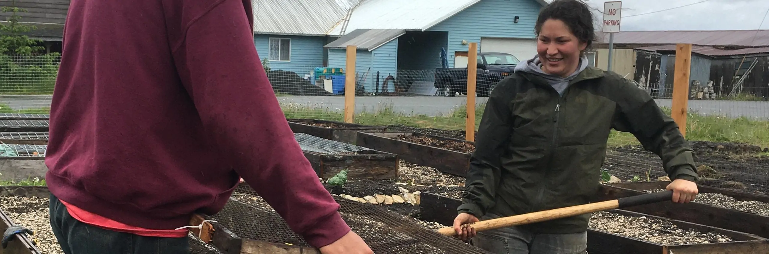 A smiling woman shovels dirt into a sift near some raised garden beds.