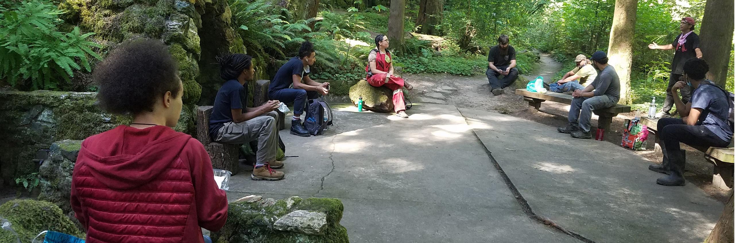 Young adults sit six feet apart. They are looking at a man standing and speaking. They are surrounded by ferns, trees, and dappled sunshine.