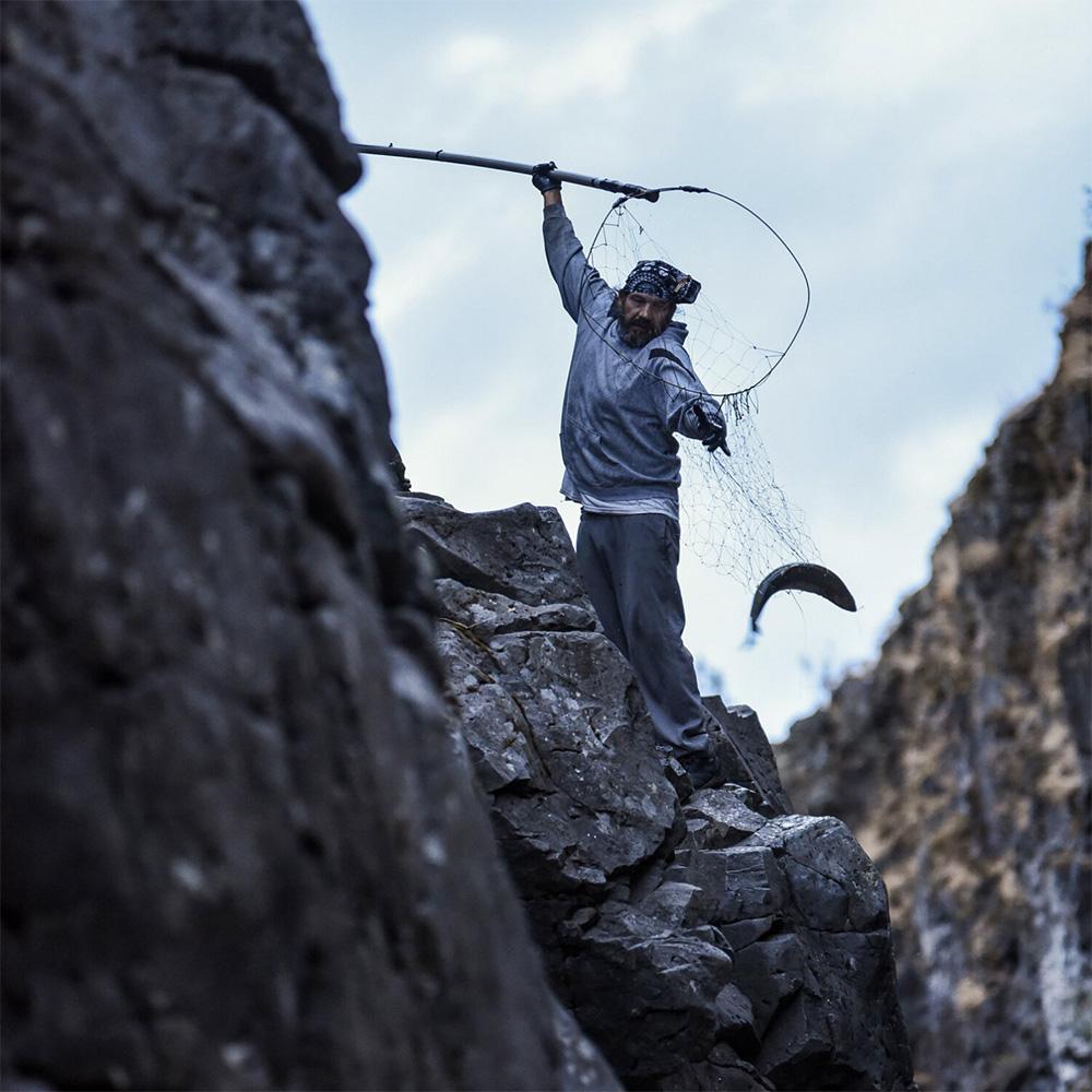 A man holds a fishing net with his right arm extended above his head. With his left arm, he reaches for a fish in the net. He's standing precariously on a rocky crag, with more crags surrounding him.