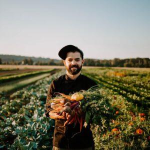 Photo of a man wearing a black cap and black chef's coat stands in a field of crops. His arms are extended toward the camera, holding an armful of harvested root vegetables.