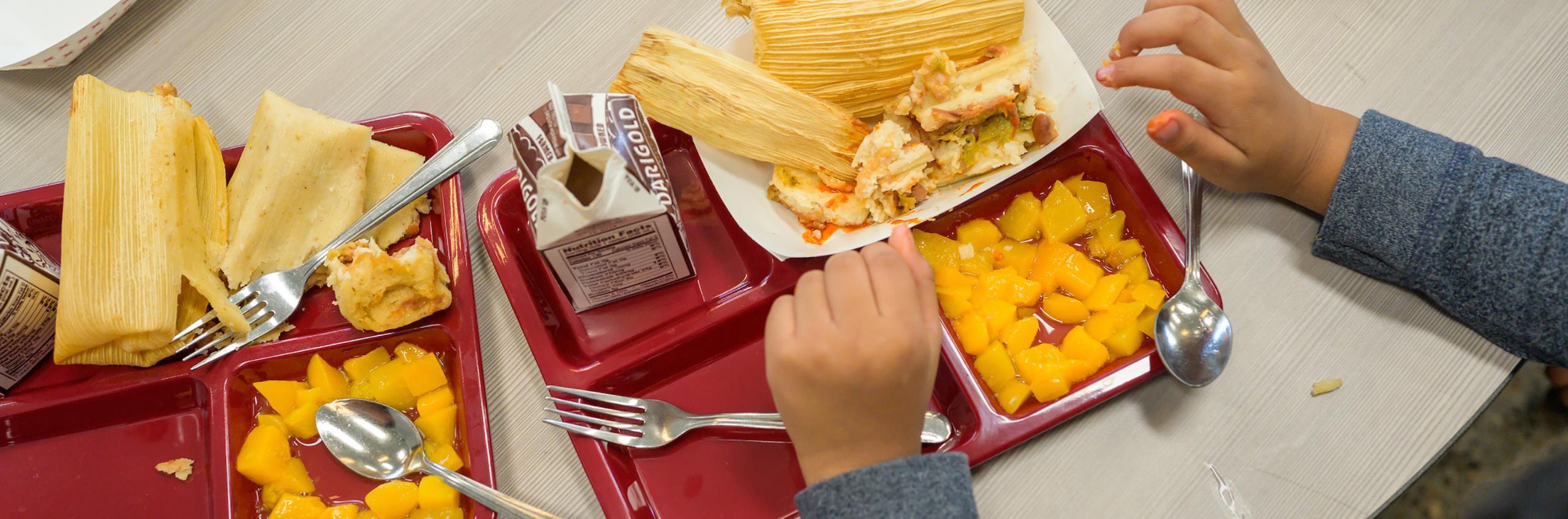 A photo taken from an overhead angle at two school lunch trays on a table. The trays hold cartons of milk, a paper tray of tamales, and canned peach chunks. A couple pairs of childrens hands are extended into the frame of the camera, resting near the trays.