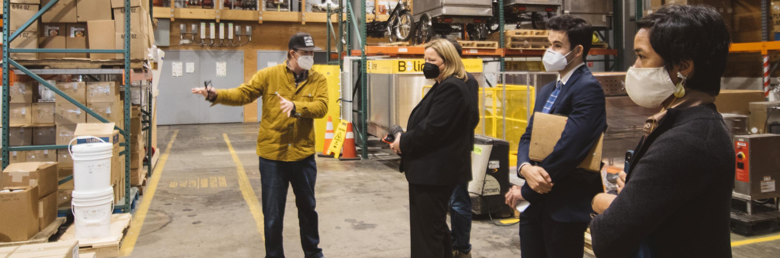 A photo of four people standing in a warehouse. The person on the left is speaking and gesturing with his hands, while the other three listen and gaze in the direction of his hands.