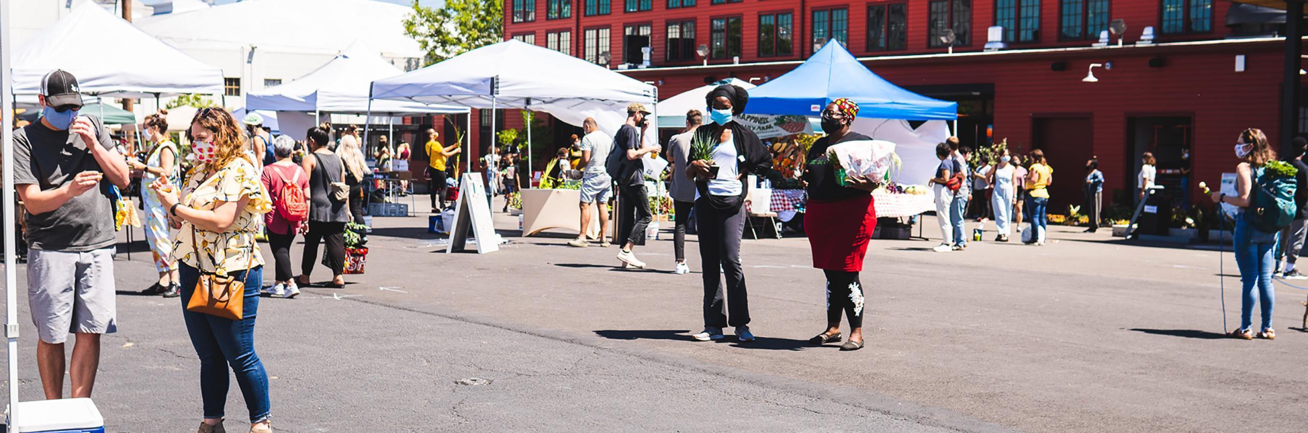 A photo of an outdoor market with customers walking around the asphalt plaza