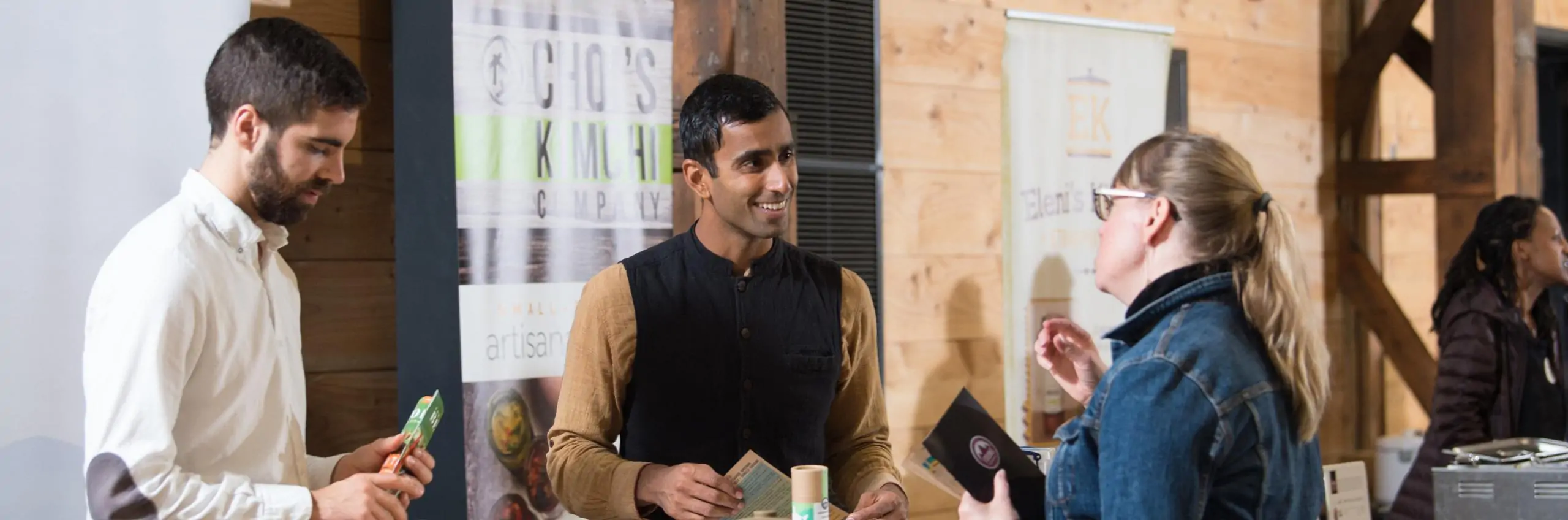 A photo of an indoor networking event, with two people standing inside a tabled booth, speaking to a customer on the other side of the table