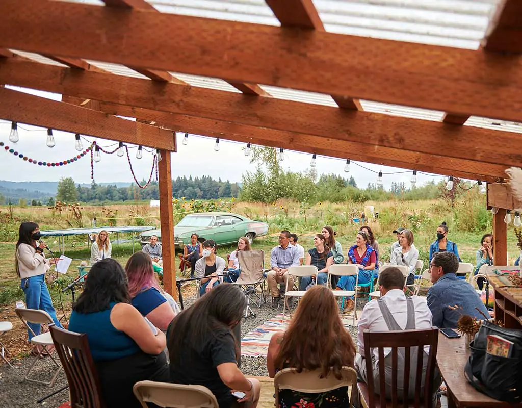 A photo of about two dozen people sitting outdoors listening to six people sitting on a platform.