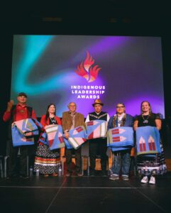 Dramatically lit stage with six people on stage on stage, backdrop screen reads Indigenous Leadership Awards