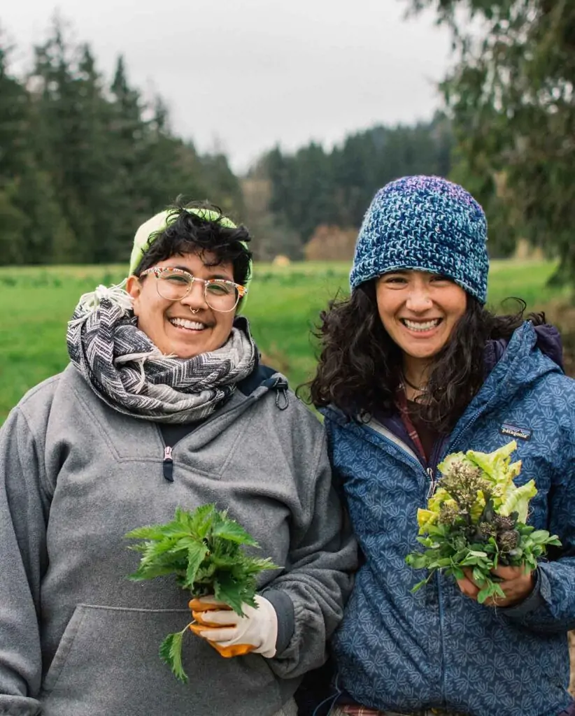 Two farmers stand side by side holding bundles of leaves