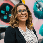 woman with glasses smiles in front of colorful mural