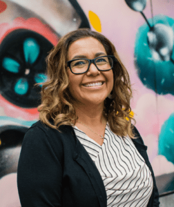 woman with glasses smiles in front of colorful mural