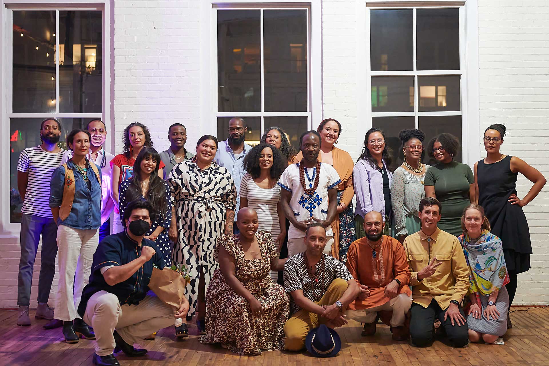 About two dozen mostly Black and Brown adults posing for a group photo, looking celebratory and joyful. They are in an indoor historic space with white brick walls and large windows.