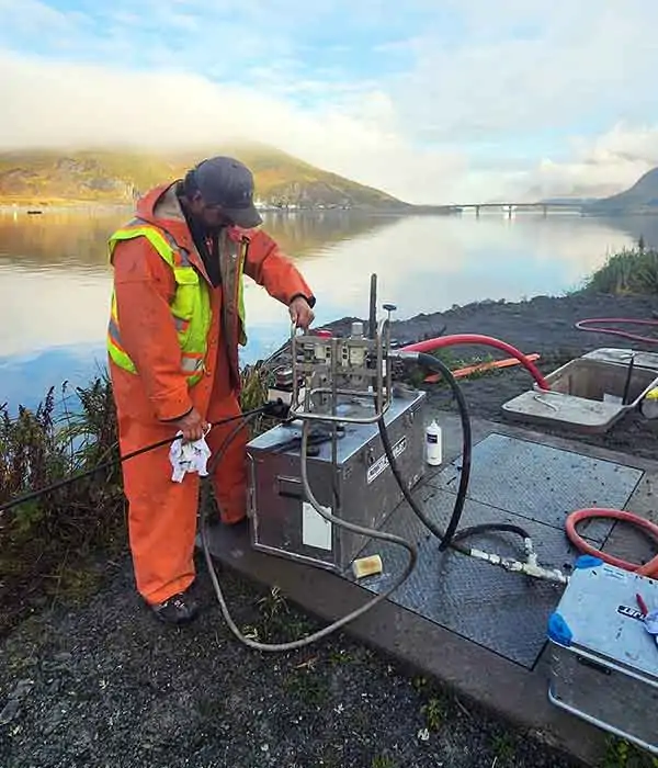 A person in a bright orange uniform and safety vest operates a large machine outdoors, next to a large calm body of water.