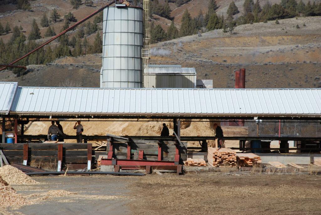 Profile of a tin-roofed, small saw mill with several people silhouetted inside and dry hills with scattered junipers in the background