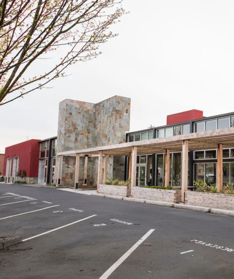 A photo of a modern, two-story office building. The exterior uses a mix of bricks, wood, large windows, and dark red paint.