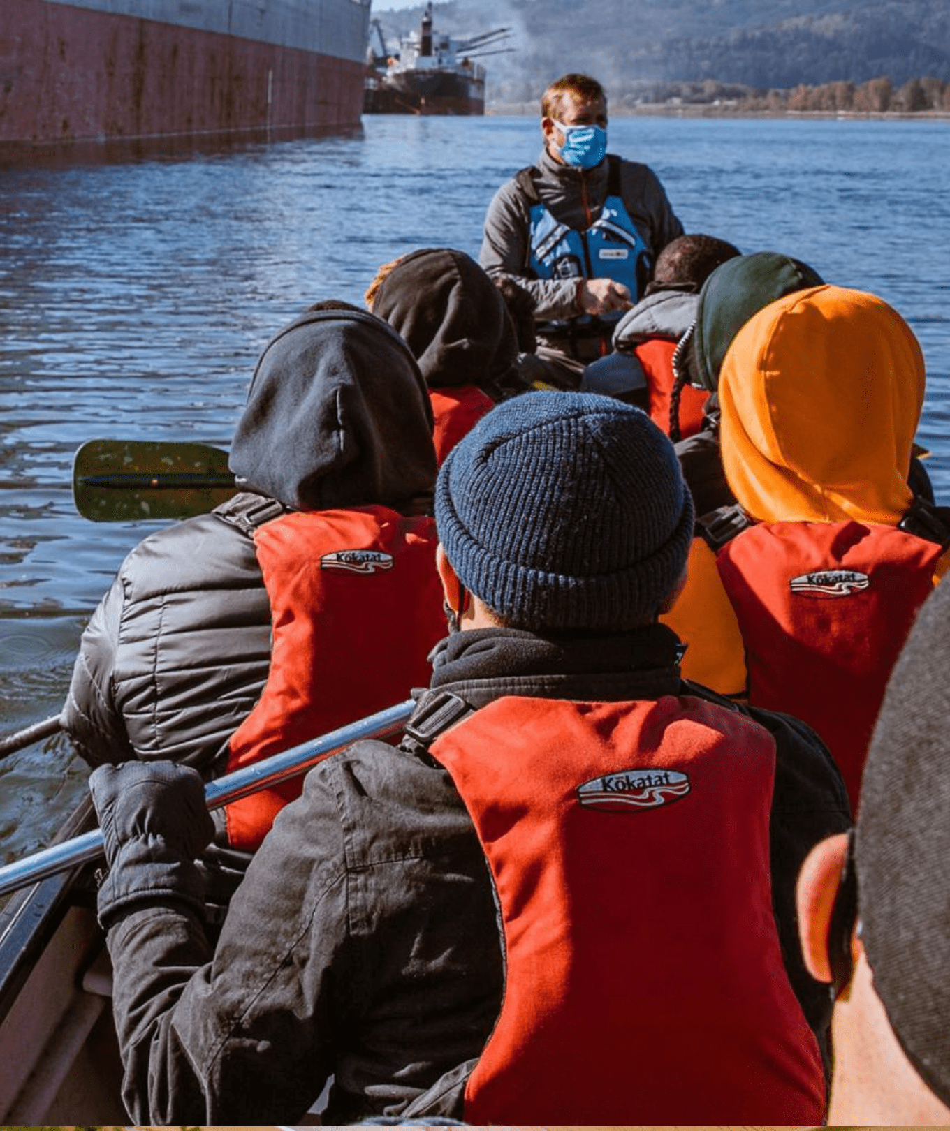 A photo taken from inside a canoe of eight people who are sitting facing away from the camera, paddling on a river. All people are wearing orange life jackets. A few people are wearing hoods or beanies on their heads.