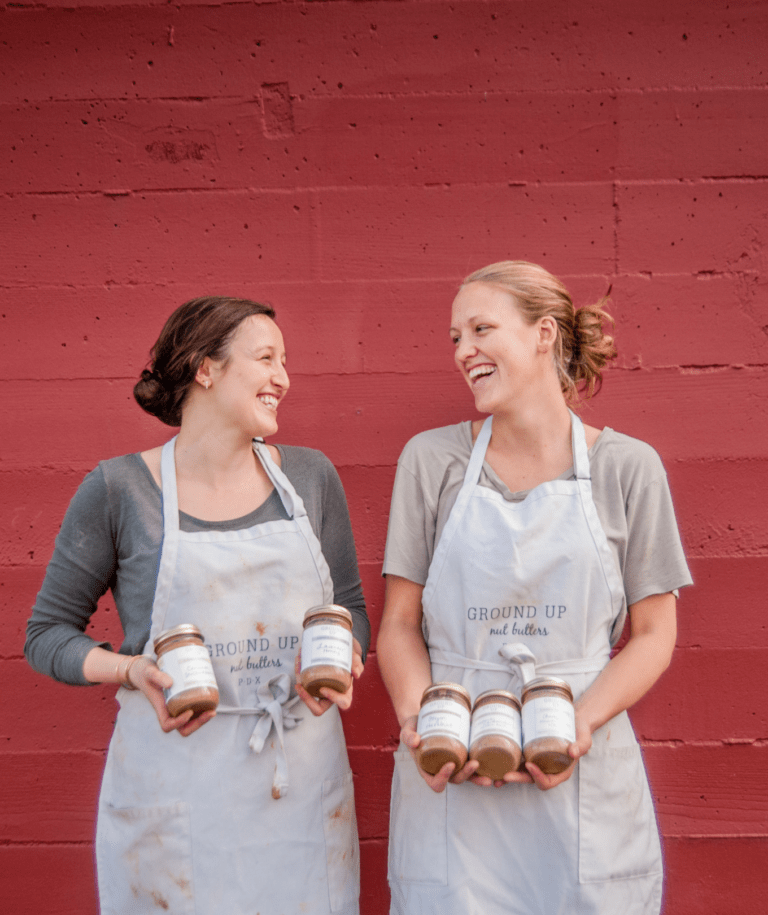 Two women, both wear aprons and holding jars of nut butter, turn toward each other with big smiles. They are standing in front of a brick wall painted red.