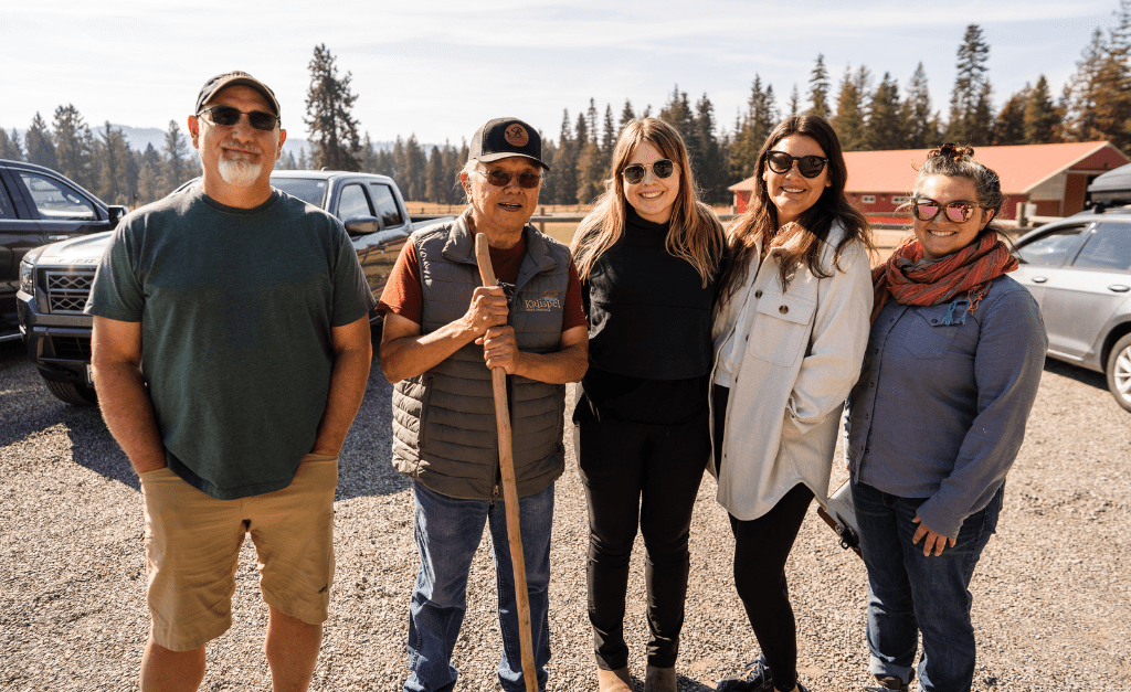 Five individuals smile and pose together for a picture in a gravel parking lot with a grassy field and trees in the background.All individuals are wearing sunglasses.