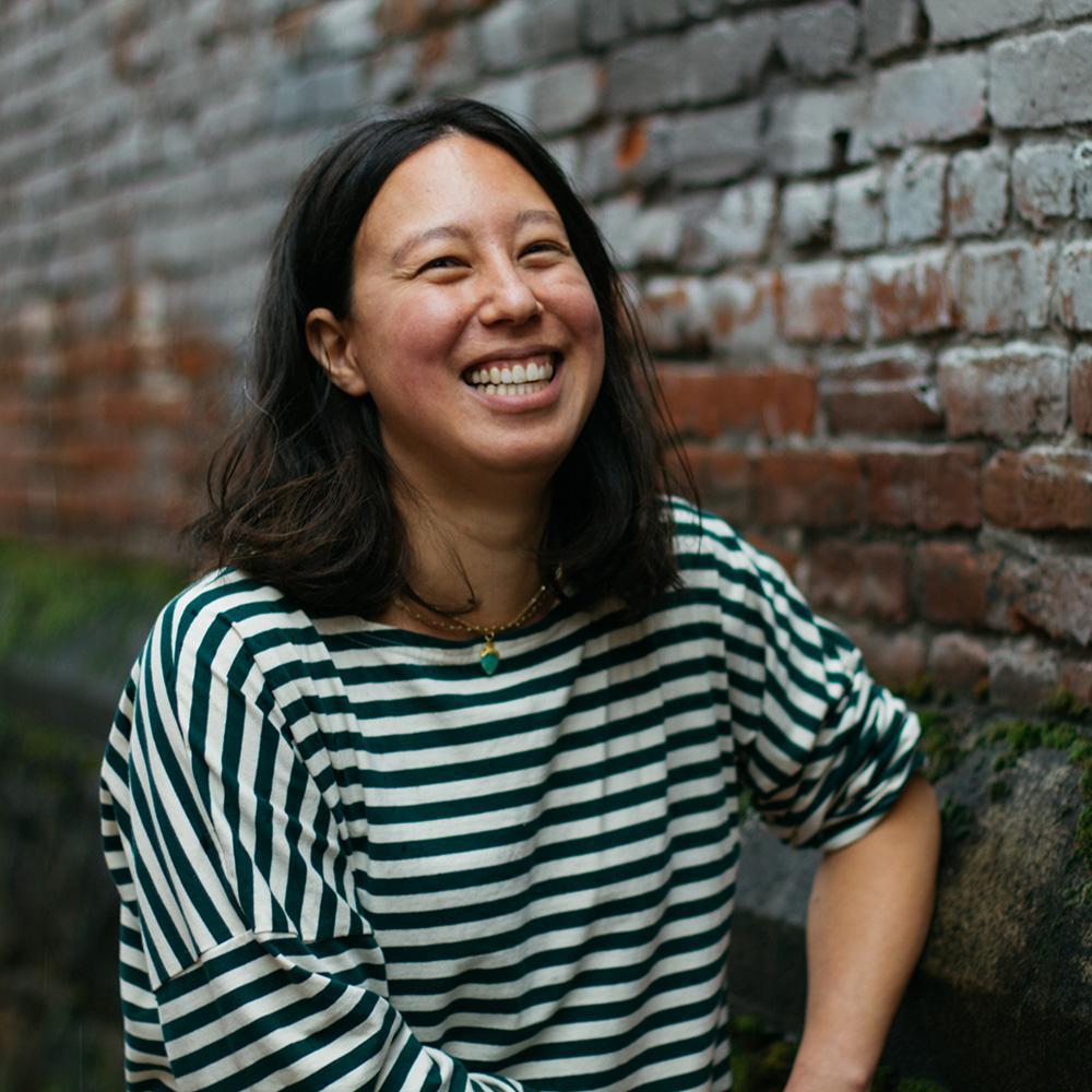 Person smiling in striped shirt, with shoulder length dark hair, brick wall background