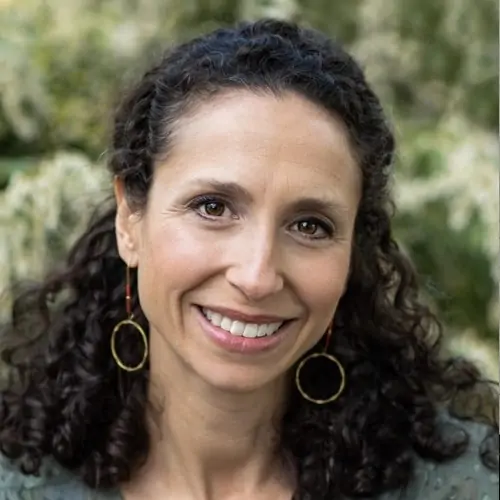 Portrait of a smiling woman with dark, curly, shoulder-length hair wearing hoop earrings and a sage green top