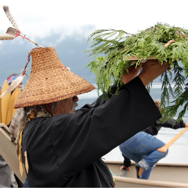A man wearing a traditional Lummi woven cedar hat and black shirt holds aloft a plank with cedar boughs
