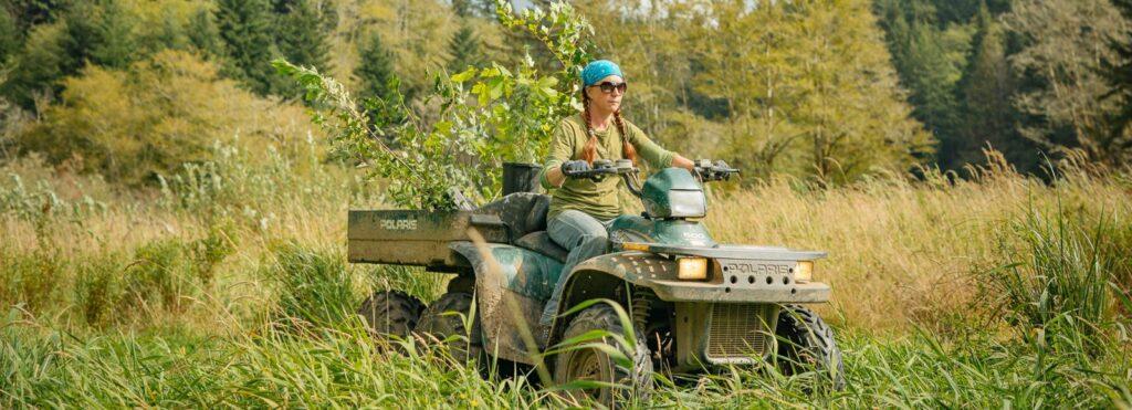 A woman in a blue bandana drives a four wheeler through tall grass with native plants in the back.