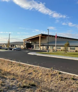 A modern-looking school facility, painted tan, with a green lawn, small parking lot, and flagpole at the front. An asphalt driveway curves from the school towards the foreground. In the background, a few clouds are scattered across a blue sky.