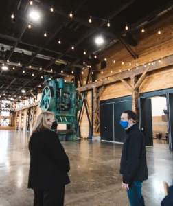 Two people, all wearing face masks, stand in a long, large, wood-paneled hall. A large green iron press can be seen in the background.