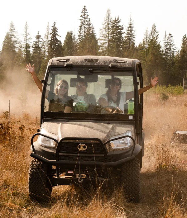 Three individuals wearing sunglasses are seated in a off-road side-by-side while driving the side-by-side through a grassy field. Two individuals weaving one of their hands outside the side-by-side for the camera. Dust is seen behind the side-by-side kicked up from driving.