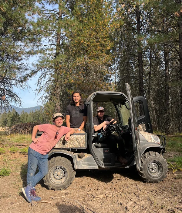 Three individuals pose for a photo around a off-road side-by-side against a forest background.