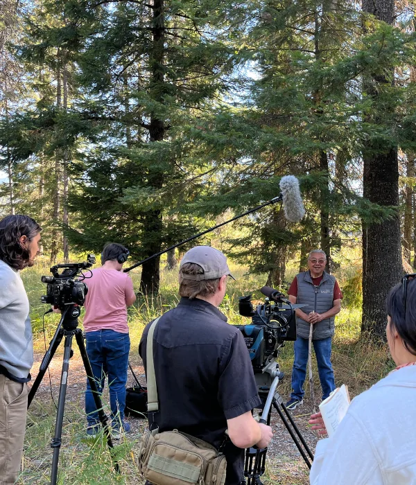 Four individuals stand with video cameras and sound equipment and face one individual who is being interviewed against the background of trees.