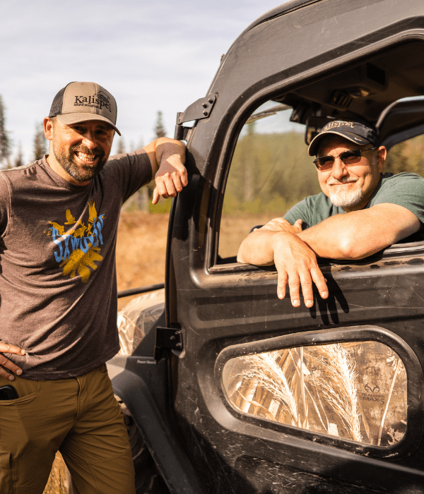 One individuals wearing a brown t-shirt, hat, and sunglasses smiles while leaning up against the door of an off road side-by-side. The second individuals smiles while seated inside the drivers seat of the off-road side-by-side with a grassy field and trees in the background.