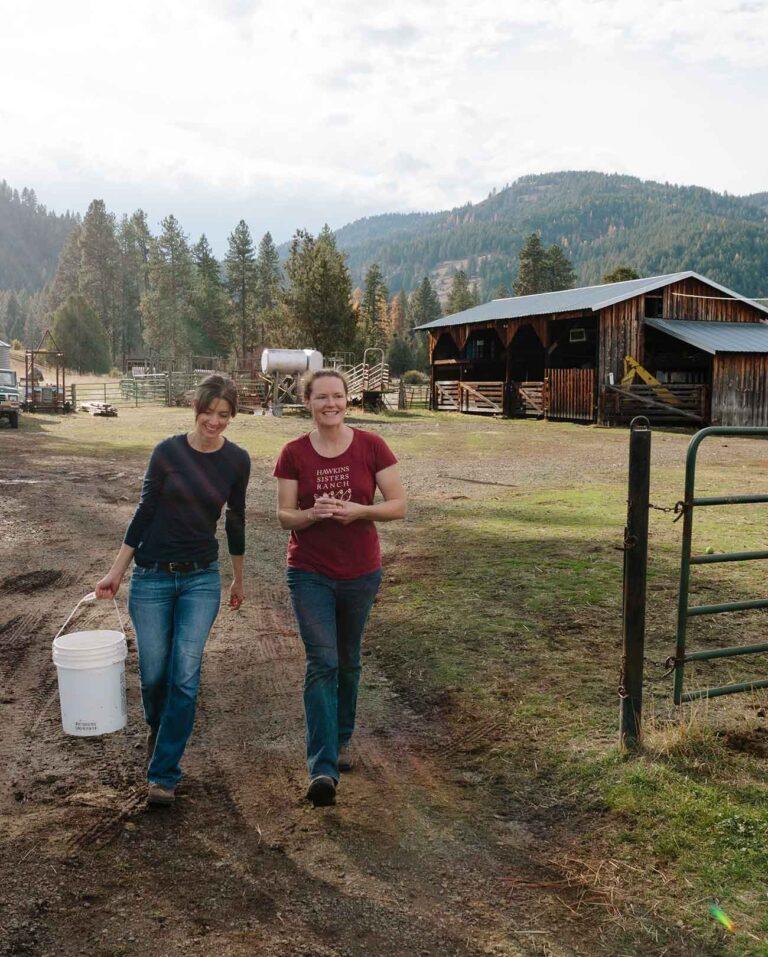 Two people walking down a dirt path in front of a farm house. One person is carrying a large five-gallon plastic bucket.