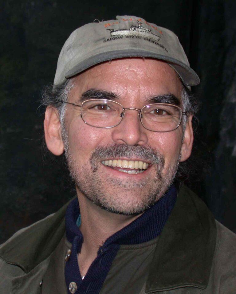 Photo of a person smiling at the camera. He wears a sage green cap and glasses and has a short salt-and-pepper beard.