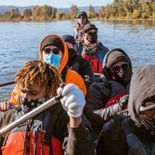 Young adults, all wearing face masks, many of whom are wearing sunglasses, beanies, or hooded jackets, row a canoe together. A tree-lined shore is about a thousand feet behind them.