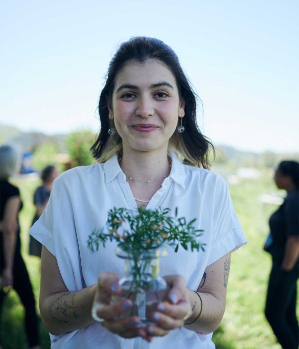 Person smiling at camera outside on a farm, holding a glass vase with rue plant in it