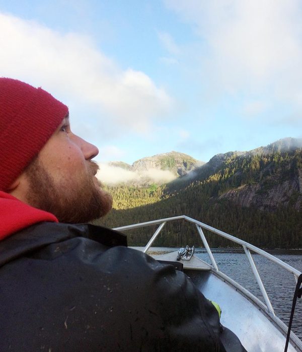 A man with a short beard and red beanie, wearing a heavy black rain coat, glances up ward at the sky. He is sitting near the bow of a small boat. He is surrounded by water and forested hills in the background.