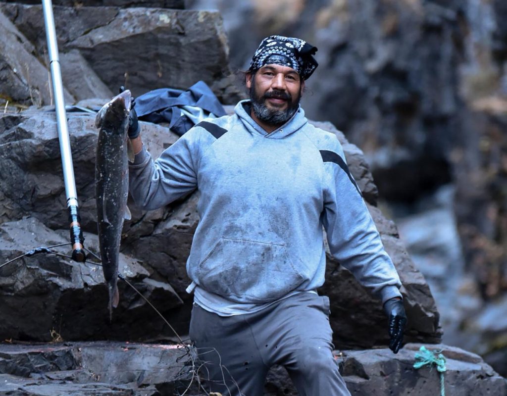 A man with a short, graying beard holds up a just-caught salmon in his right hand, with a slight smile toward the camera. He is wearing a gray hoodie and black bandana on his head. He's surrounded by rocky crags.