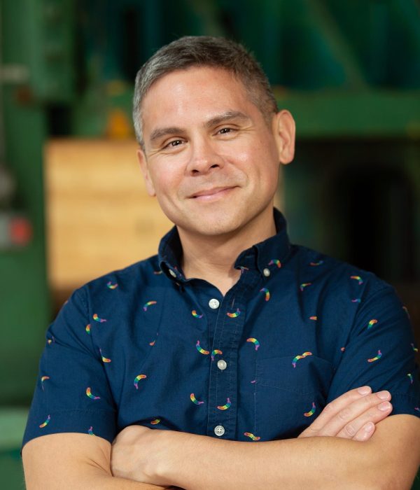 A photo of a person with his arms crossed, smiling facing the camera. He has short hair and wears a dark blue collared button-up shirt, with a print of small rainbow motifs.