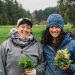 Two farmers stand side by side holding bundles of leaves