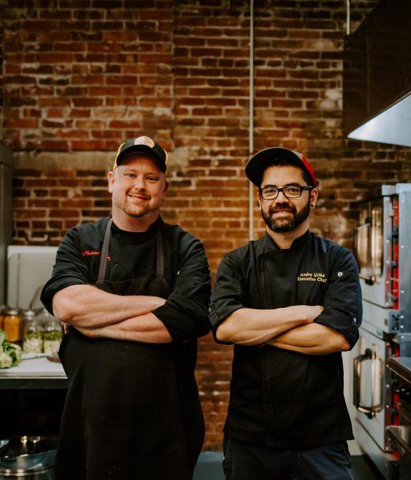A photo off two men posing with their arms crossed and smiling to the camera. They are wearing black caps and black chef uniforms. They are standing in an industrial kitchen with a brick wall.