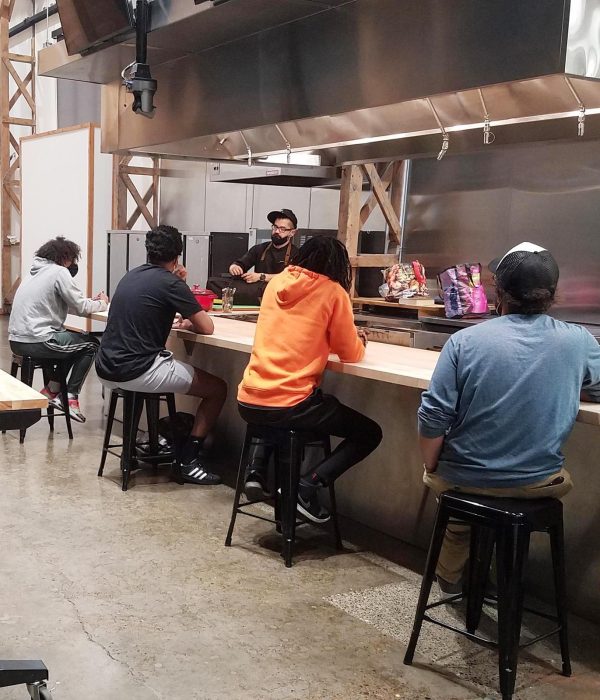 A photo of four young people sitting on stools along an industrial kitchen counter. On the other side of the counter, a man wearing a black cap and black chef's uniform is speaking while gesturing with his hands.