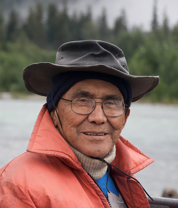 Portrait of an older man wearing glasses, a bucket hat fitted over his beanie, and an orange rain jacket over a knitted sweater