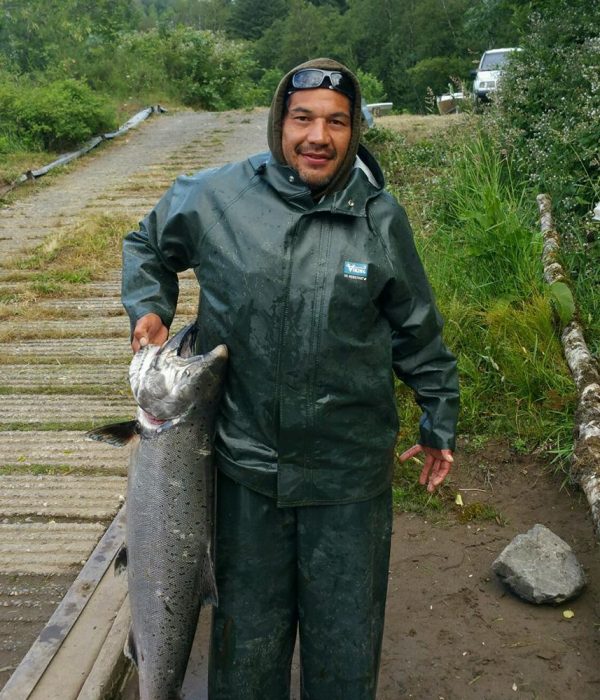 A man wearing a green rain jacket holds up a large, freshly caught salmon that is almost half the length of his standing height. He is standing near a boat ramp leading up to a parking lot just barely visible in the background.