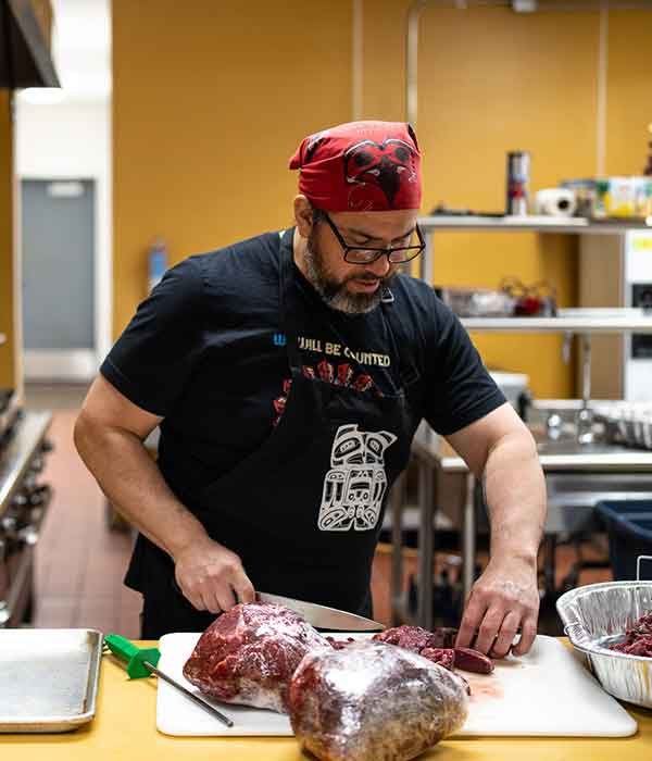 A person wearing a red bandana and glasses prepares deer meat in a community center's kitchen