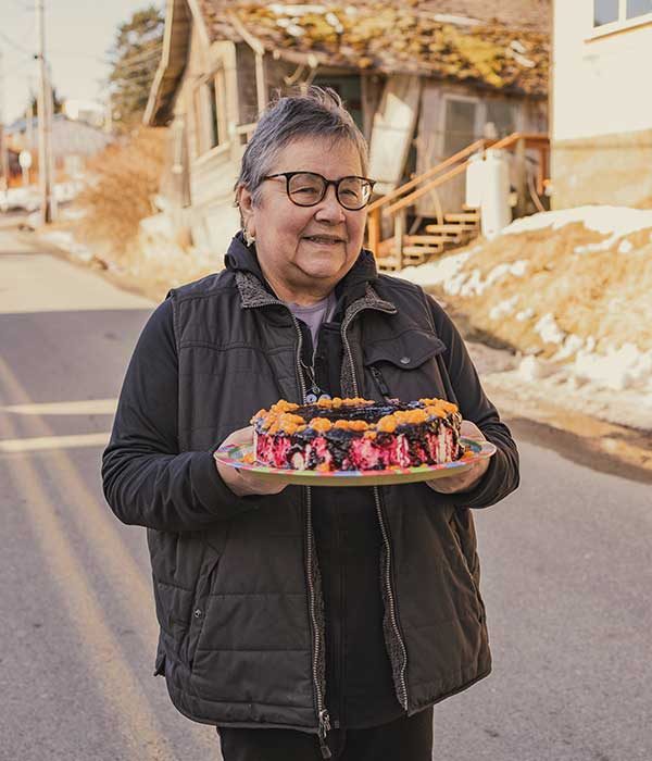 An older woman with short gray hair, wearing glasses and a vest, holds a plate of homemade cloudberry cheesecake that is dripping with a dark berry sauce.