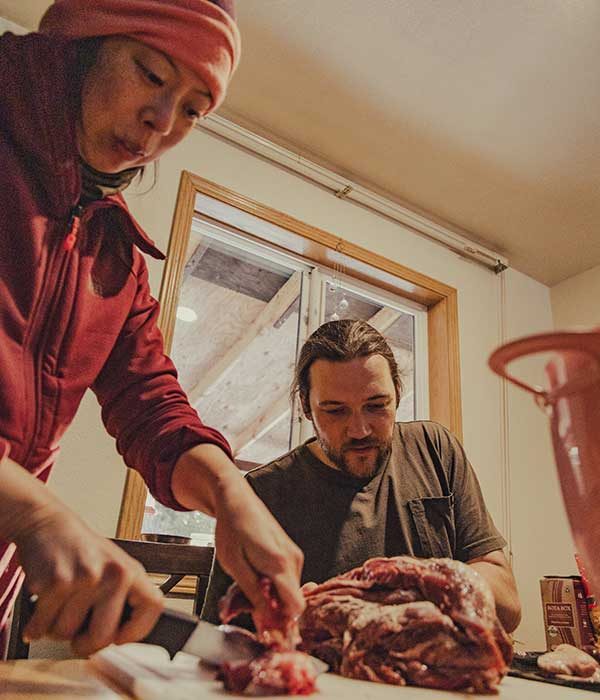 Two people in a home kitchen, cutting raw deer meat on a cutting board