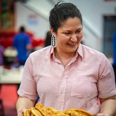 A picture of a person wearing beaded earrings and a pink striped button-up shirt carries a box of fresh frybread