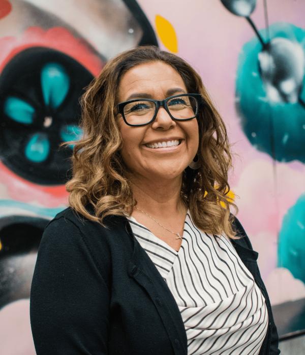 woman with glasses smiles in front of colorful mural