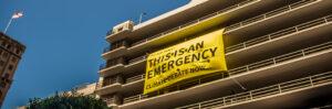 A photo of a banner that says "This is an Emergency, climate debate now" hung over a parking structure