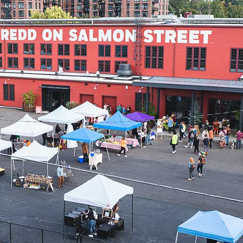 aerial view of a farmers market, grouping of tents, sunny day, red building in background
